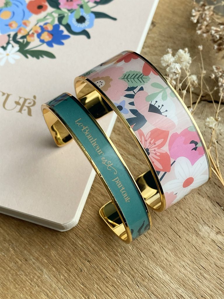 Deux bracelets joncs élégants avec motifs floraux colorés et inscriptions dorées posés sur une surface en bois, à côté d'une carte avec des fleurs