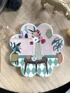 Sous-verre en céramique en forme de fleur avec une illustration colorée de fleurs dans un vase sur une table, sur fond pastel avec des motifs géométriques et des plantes en pot, finition brillante et dos antidérapant en liège